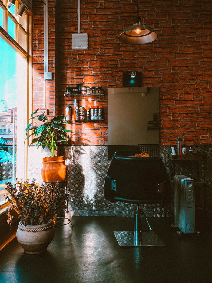 why-choose-us-02 Modern barbershop interior featuring a chair, plants, and brick wall design with natural daylight.